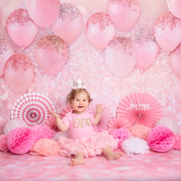 One-year-old baby girl photographed on pink balloon backdrop during birthday milestone session