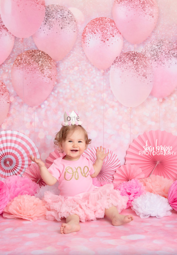 One-year-old baby girl photographed on pink balloon backdrop during birthday milestone session