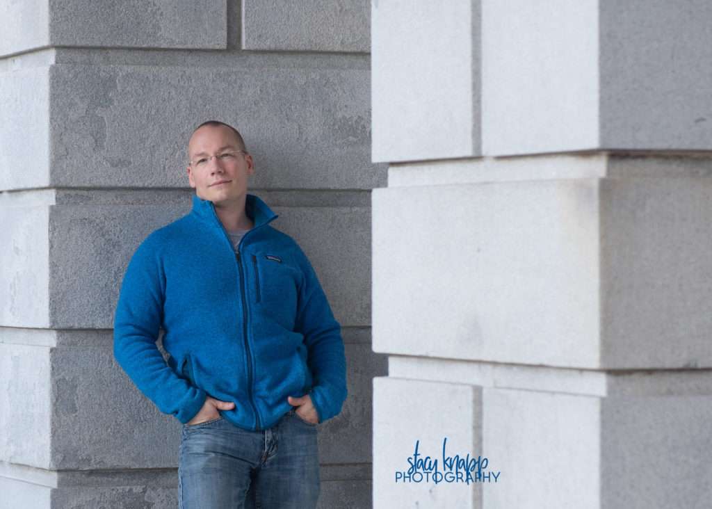 Headshot of photograph a man standing in front of the State House in Augusta Maine wearing blue