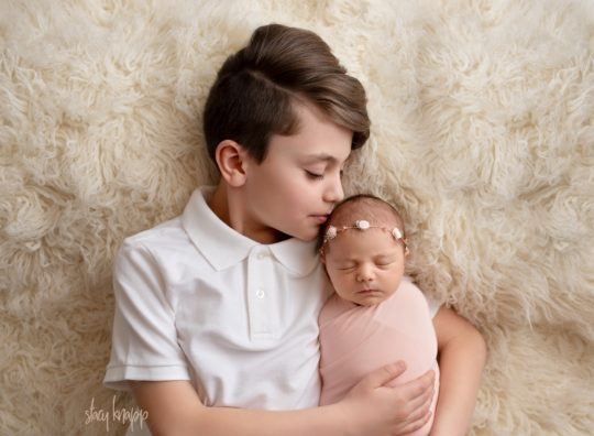 This is a photo of a Maine newborn baby girl and her big brother taken by Maine photographer Stacy Knapp Photography. The newborn baby girl is wearing a pink wrap and wearing a headband. The older sibling is kissing baby on the head.
