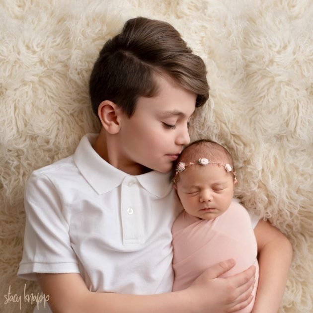 This is a photo of a Maine newborn baby girl and her big brother taken by Maine photographer Stacy Knapp Photography. The newborn baby girl is wearing a pink wrap and wearing a headband. The older sibling is kissing baby on the head.