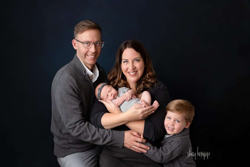 Photo of Maine newborn family with mama holding a newborn baby alongside her son and the baby's father. Family is wearing gray and they're on a navy blue backdrop.