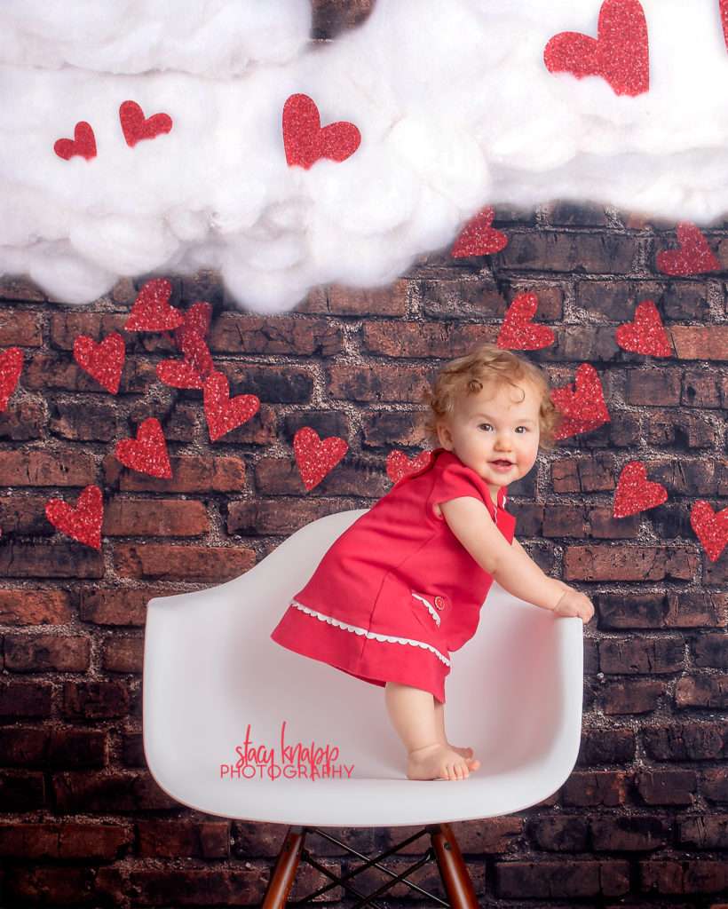 Baby girl photographed on Valentines Day backdrop with hearts and clouds during mini session