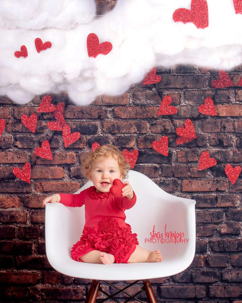 Baby girl photographed on Valentines Day backdrop with hearts and clouds during mini session