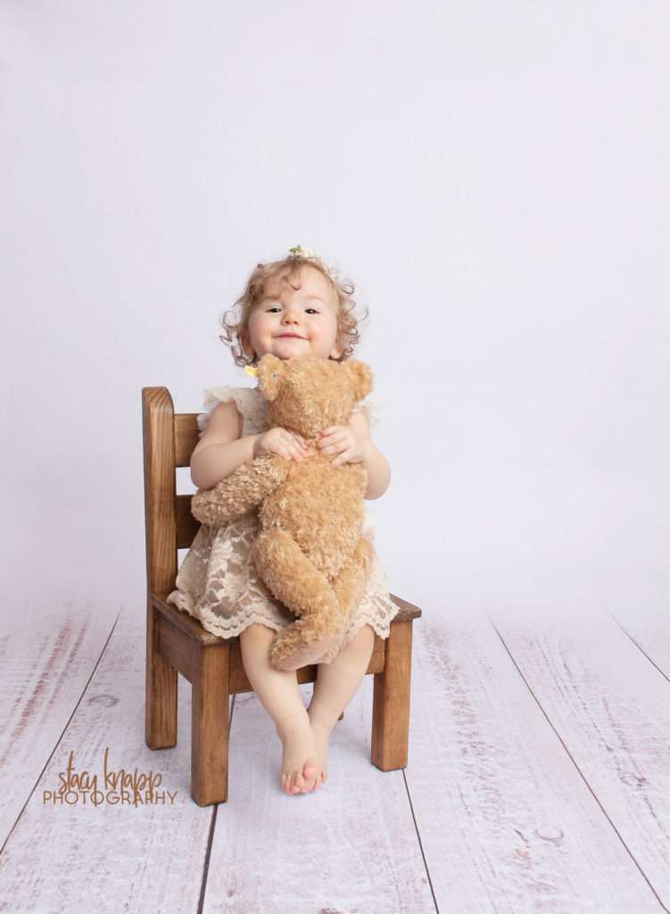 Photo of toddler girl in beige dress on white wood backdrop in sitter chair with teddy bear