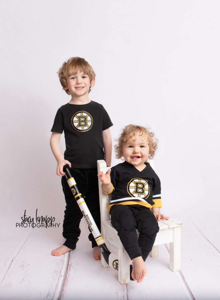 Photo of preschool boy and toddler girl in Bruins outfits with hockey stick on white wood backdrop