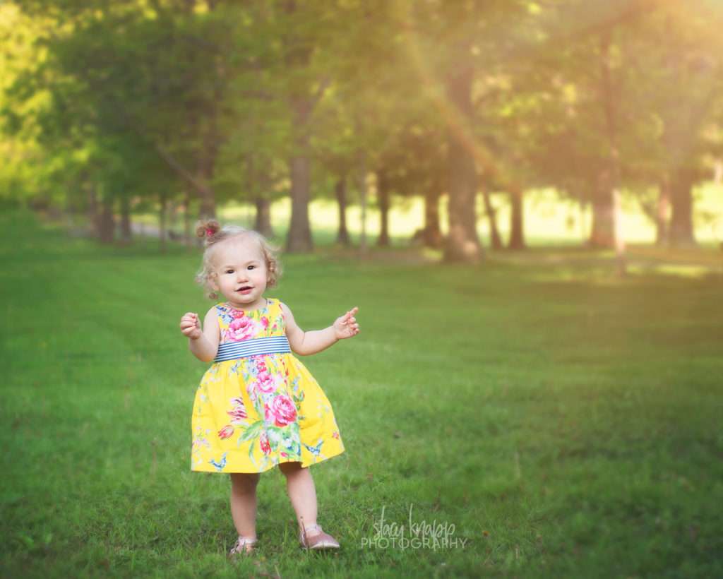 Toddler girl in Capital Park in Augusta, Maine walking in grass wearing yellow floral dress