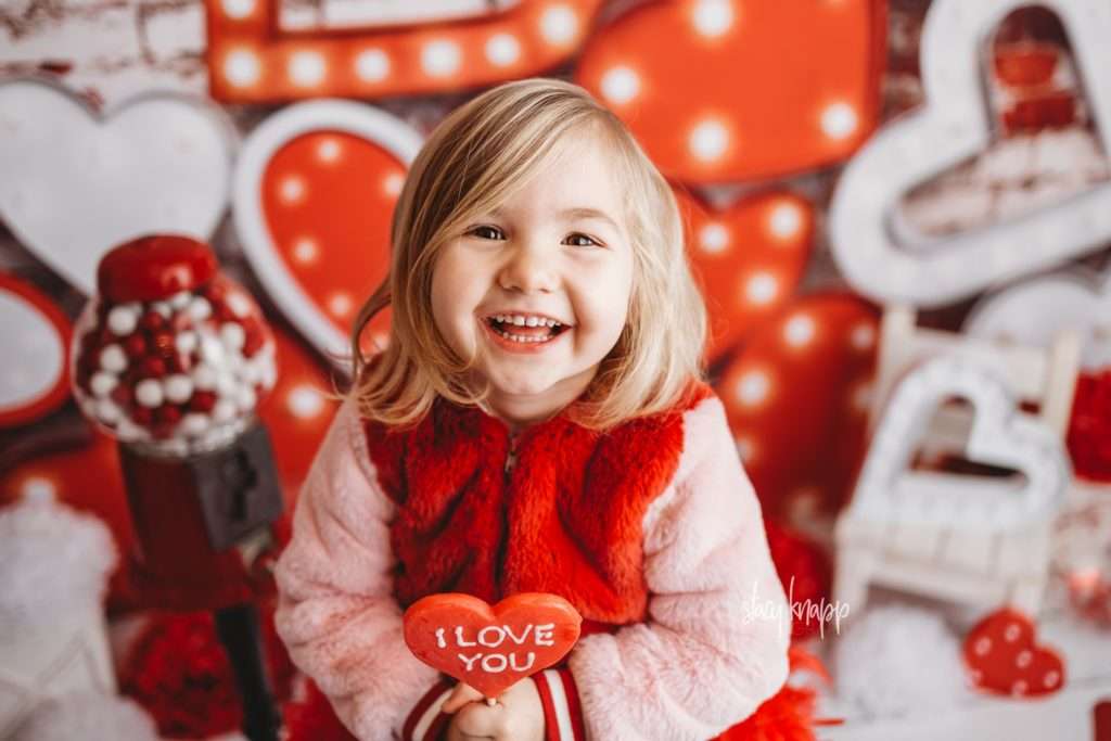Young girl celebrating valentines day at Stacy Knapp Photography holding heart lollipop