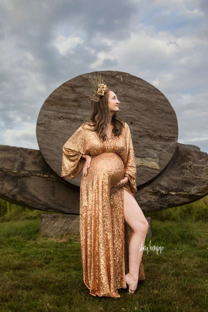Maternity photo of a pregnant mother in a gold glittery gown wearing a crown in front of stone art at the Viles Arboretum in Augusta, Maine by photographer Stacy Knapp Photography