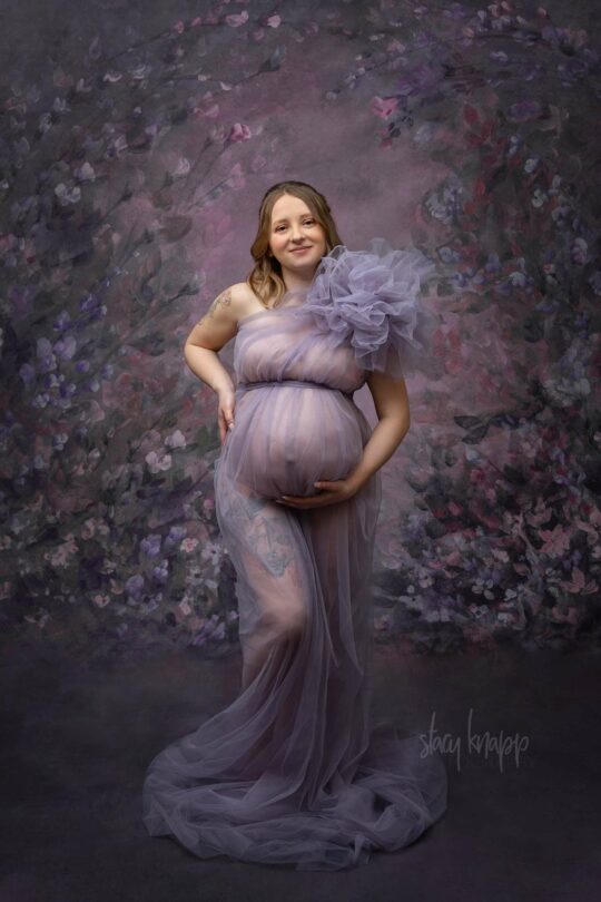 This is a maternity photo of a pregnant Maine mother in a purple gown with a floral background by photographer Stacy Knapp Photography