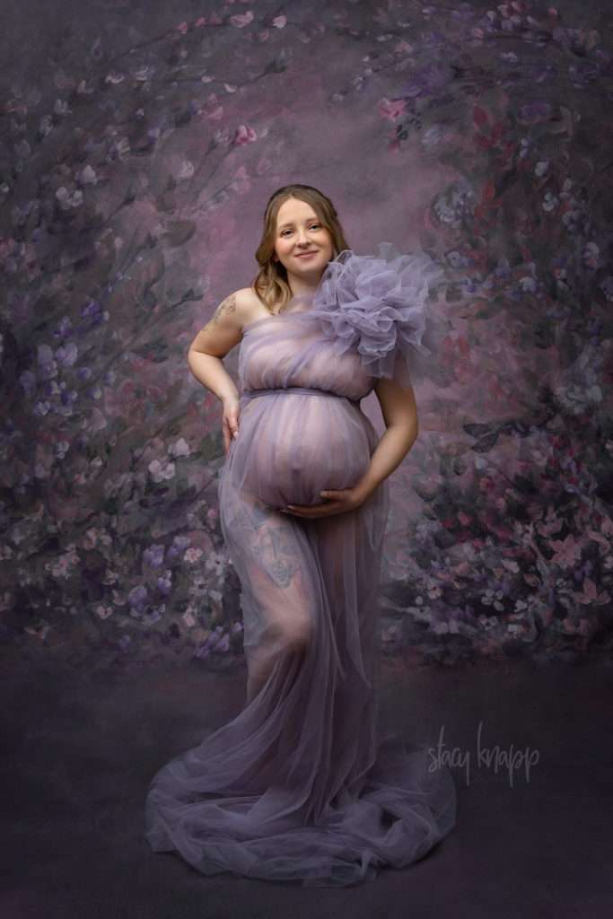 This is a maternity photo of a pregnant Maine mother in a purple gown with a floral background by photographer Stacy Knapp Photography