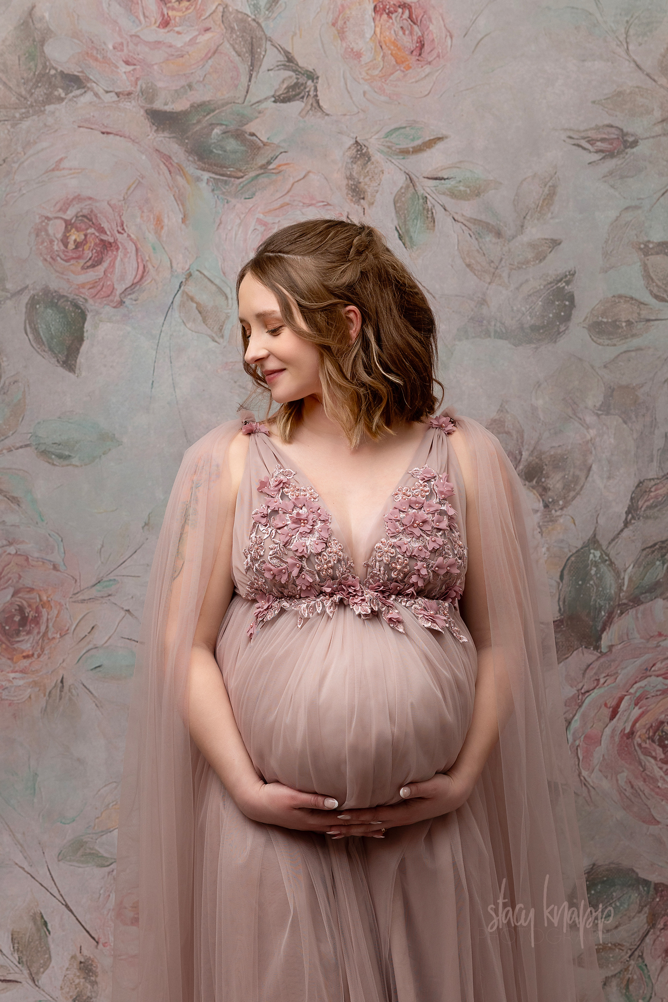 Close-up maternity photo of a pregnant Maine mother in a pink floral gown with a pink background by photographer Stacy Knapp Photography