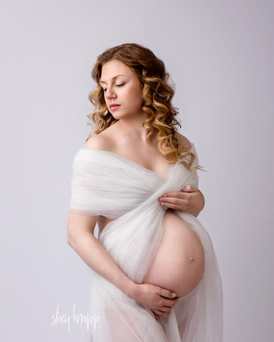 This is a maternity photo of a pregnant Maine mother wrapped in white tulle fabric with a white background by photographer Stacy Knapp Photography