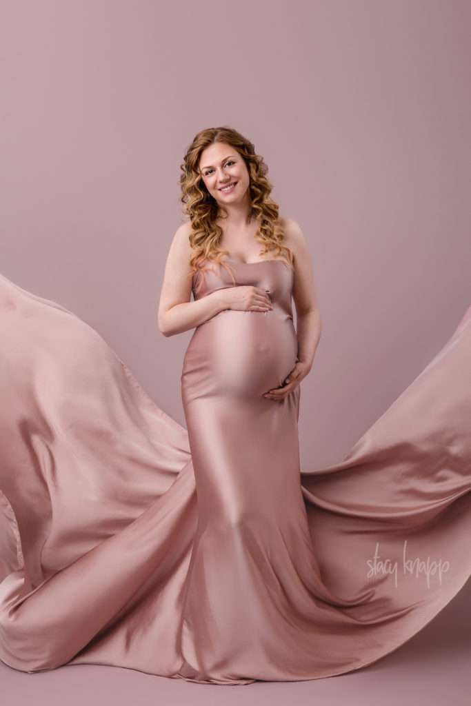 Maternity photo of a pregnant Maine mother wrapped in pink fabric as a gown with flowing fabric sides on a pink background by photographer Stacy Knapp Photography