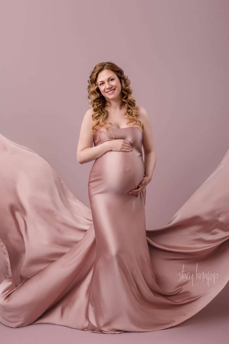 Maternity photo of a pregnant Maine mother wrapped in pink fabric as a gown with flowing fabric sides on a pink background by photographer Stacy Knapp Photography