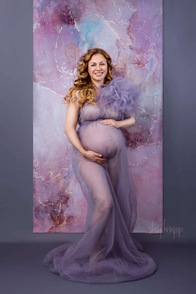 Maternity photo of a pregnant Maine mother in a purple gown with a gray and printed layered background by photographer Stacy Knapp Photography