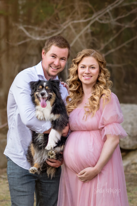 This is a maternity photo of a pregnant Maine mother in a pink gown with her husband and family dog taken outside their home by photographer Stacy Knapp Photography