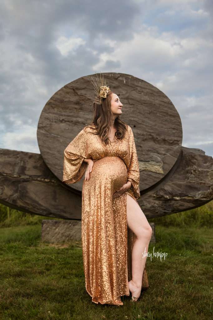 Maine maternity photo of pregnant mother in gold glitter gown in front of a stone sculpture in the Viles Arboretum in Augusta by photographer Stacy Knapp Photography
