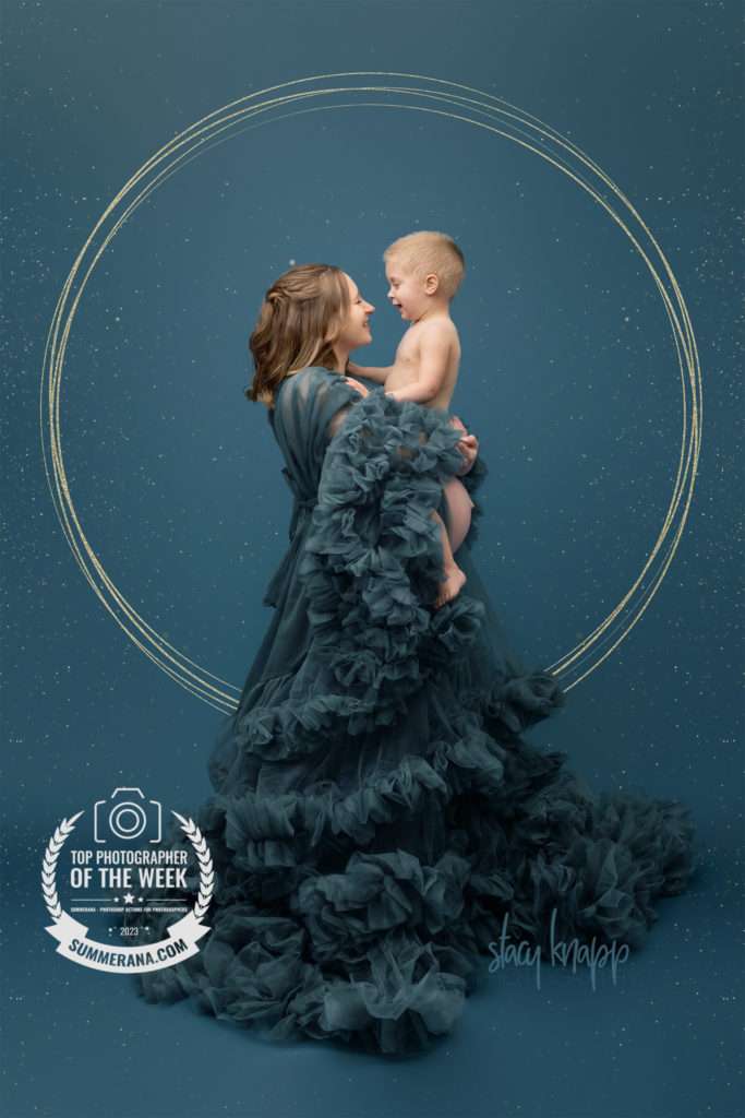 Photo of a young mother wearing a teal luxury maternity gown holding her toddler baby boy while pregnant with a baby girl.