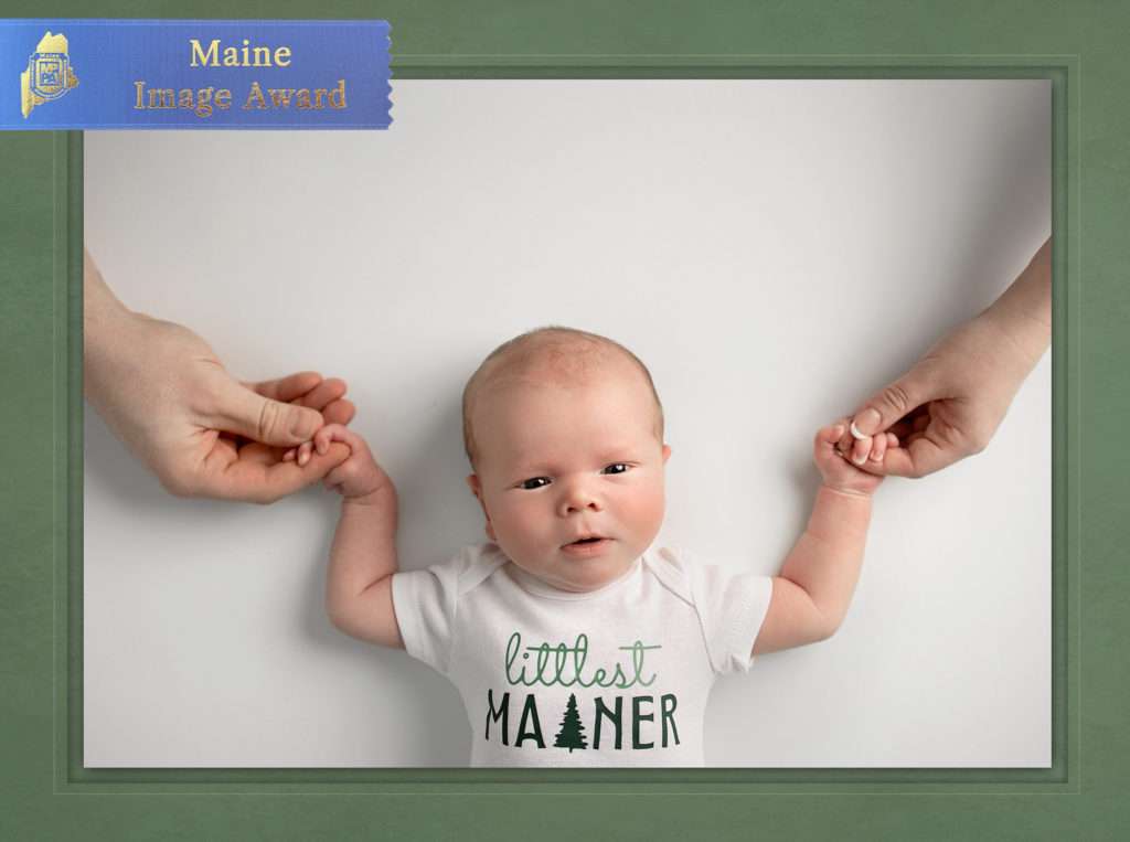 MPPA Maine blue ribbon merit award winning photo of Maine newborn baby boy wearing blue with his parents hands holding him by Stacy Knapp Photography